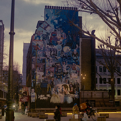 The image features a large building with a mural on its side, which is covered in many pictures. There are several people walking around the area, some of them carrying backpacks and handbags. A few bicycles can be seen parked nearby, indicating that this might be a popular spot for pedestrians to visit or pass through.