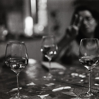 The image is a black and white photo of a woman sitting at a table with two wine glasses in front of her. She appears to be making a peace sign gesture, possibly indicating that she's enjoying the wine or having a good time. There are also two bottles on the table, one near the left edge and another towards the right side.
