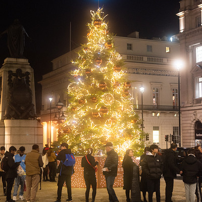 The image features a large, lit-up Christmas tree in the center of a city square. A group of people is gathered around the tree, admiring its beauty and enjoying the festive atmosphere. There are at least 13 individuals visible in the scene, with some standing closer to the tree while others are further away.