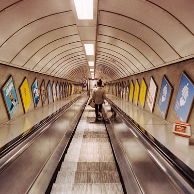 The image depicts a busy subway station with people walking on the escalator. There are several individuals visible, some of them carrying handbags and backpacks. A person is standing at the top of the escalator, possibly waiting for others to join them or preparing to exit the station.