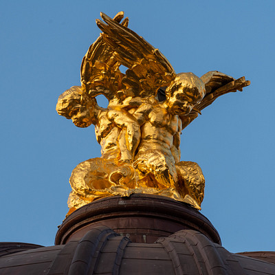 The image features a large, ornate statue of an angel or mythical creature with gold wings. It is situated on top of a building, possibly a church steeple, and appears to be made of metal. The statue has a prominent position in the scene, drawing attention due to its size and intricate design.