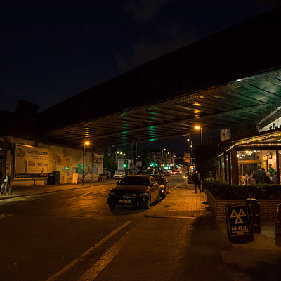 The image depicts a night scene with a car driving down an empty street under a bridge. There are several people walking around the area, some of them carrying handbags. A few cars can be seen parked or moving along the road.