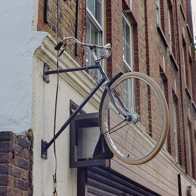 The image features a bicycle parked on the side of a building, hanging from a hook or a metal bracket. It appears to be an old-fashioned bike with a basket attached to it. The scene is set in front of a brick building, and there are two potted plants visible in the background.