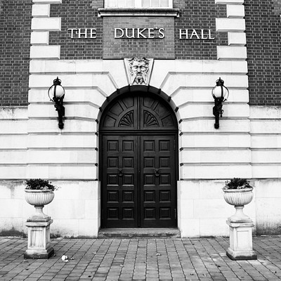 The image features a black and white photo of an old building with a large arched doorway. Above the door, there is a sign that reads "The Duke's Hall." Two potted plants are placed on either side of the entrance, adding to the overall charm of the scene. In addition to the main entrance, there are two smaller doors visible in the image, one located towards the left and another further back on the right side.