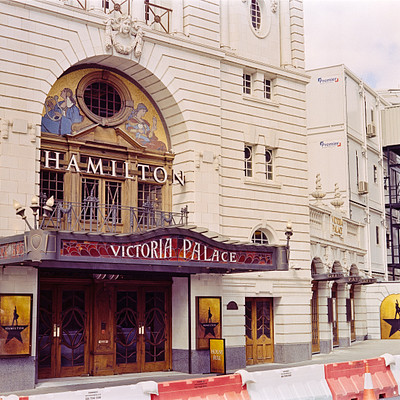 The image features a large, ornate theater building with a prominent sign that reads "Hamilton Victoria Palace." The theater is located on a street corner and has an old-fashioned appearance. There are several statues adorning the facade of the building, adding to its grandeur. In addition to the main theater entrance, there are two other doors visible in the scene.