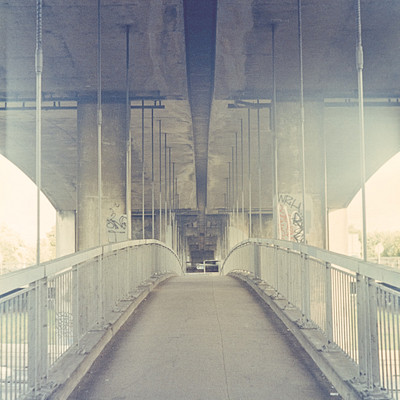 The image is a black and white photo of an empty bridge with a train passing underneath. There are several cables hanging from the ceiling, creating a unique atmosphere. A car can be seen on the bridge, traveling across it.