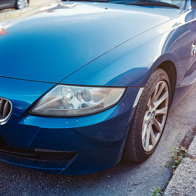 The image features a blue sports car parked on the side of a road. The car is positioned in such a way that it takes up most of the frame, with its hood and roof visible. There are two people standing near the car, one closer to the left side and another further back on the right side.