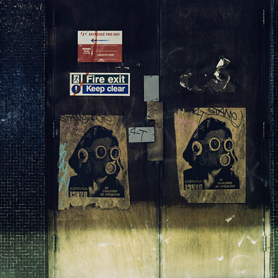 The image features a doorway with two signs on it. One sign is a fire exit sign, while the other has a picture of a gas mask and says "keep clear." There are also several stickers on the door, adding to its unique appearance. The scene appears to be in black and white, giving it an artistic touch.