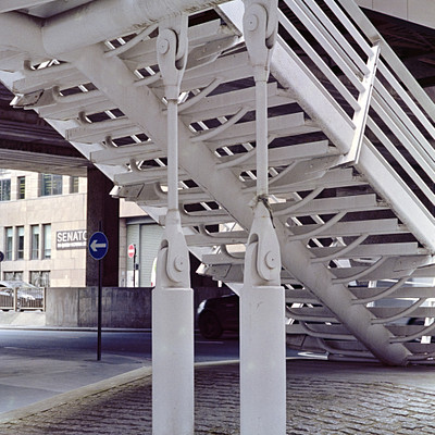 The image features a white staircase with metal railings, located under an overpass. There are two sets of stairs in the scene, one on the left side and another on the right side. Above these stairs, there is a bridge that provides shelter from the elements.