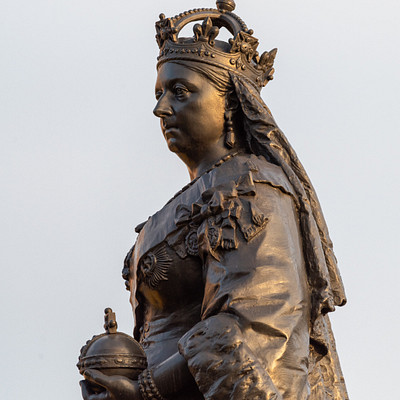 The image features a statue of a woman wearing a crown, standing in front of the sky. She is holding a small ball or sphere in her hand. The statue appears to be made of metal and has an ornate design, giving it a regal appearance.