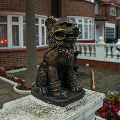 The image features a large, intricately carved statue of a lion sitting on top of a stone pedestal. The lion statue is positioned in front of a house with red brick walls and white trim. There are several potted plants surrounding the area, adding to the overall ambiance of the scene.