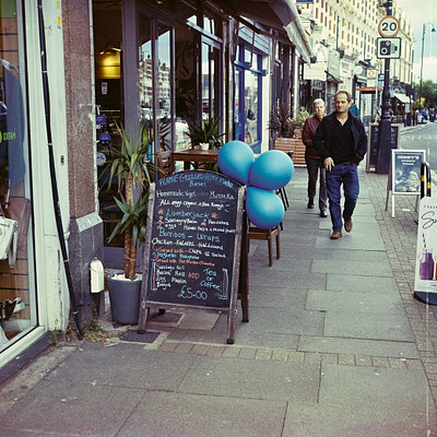 The image is a black and white photo of a man walking down the sidewalk in front of a store. There are several potted plants placed along the sidewalk, with one near the center of the scene and two others closer to the right edge. A chalkboard menu sign can be seen on the left side of the image, likely advertising food or drinks from the nearby store.