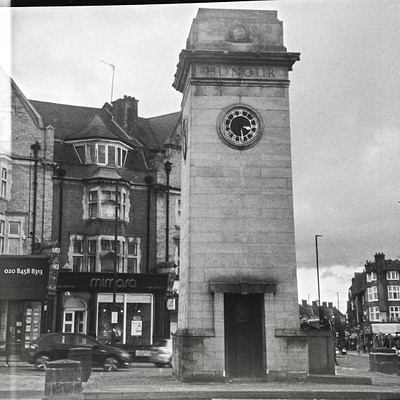 The image is a black and white photo of an old clock tower in the middle of a city. The clock tower stands tall, with its face visible on all sides. It appears to be located near some buildings, possibly a church or a historical site.