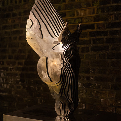 The image features a unique sculpture of an animal, possibly a horse or a bird, made from metal. It is displayed on a pedestal in front of a brick wall, which adds to the artistic atmosphere of the scene. The sculpture appears to be a focal point and draws attention due to its intricate design and craftsmanship.