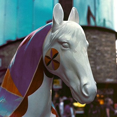 The image features a large, colorful horse statue with a heart-shaped design on its forehead. This unique and eye-catching sculpture is located in the middle of a busy street, surrounded by people walking around it. There are at least 12 individuals visible in the scene, some closer to the horse while others are further away.