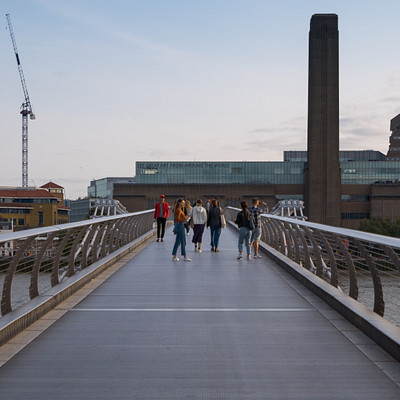 The image features a group of people walking across a bridge over water. There are at least 12 individuals visible in the scene, some closer to the foreground and others further away. They appear to be enjoying their walk on the bridge, which is situated above a river.