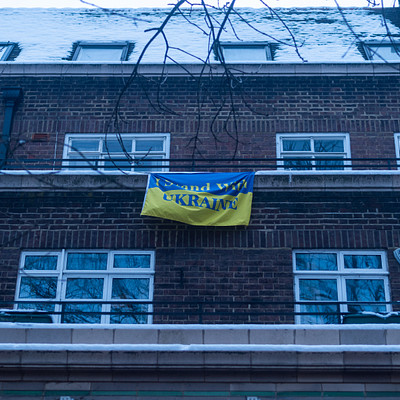 The image features a building with a blue and yellow flag hanging from the side of it. The flag is positioned above a window, making it stand out against the brick wall. There are several windows on the building, some located near the top while others are situated at various heights throughout the structure.