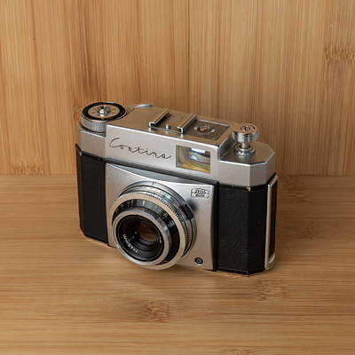 The image features a vintage camera sitting on top of a wooden table. The camera is an old-fashioned model, possibly a Kodak, and it has a black case with silver accents. It appears to be well-maintained and ready for use.