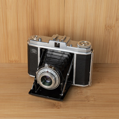 The image features an old-fashioned camera sitting on a wooden table. The camera is black and silver, with its lens facing the viewer. It appears to be an antique or vintage model, possibly from the 1950s era.