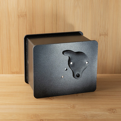 The image features a small black box with a handle on top of it. The box is placed on a wooden table, which appears to be made from wood planks. The box has a unique design and seems to be an interesting piece of decor or functional item for the space where it's located.