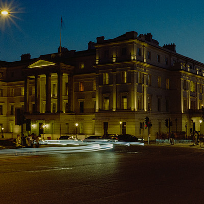 The image is a black and white photo of a large, stately building with columns in front. There are several cars parked outside the building, including one on the left side, two in the middle, and another on the right side. A traffic light can be seen near the center of the scene.