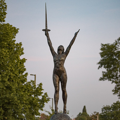The image features a statue of a woman holding a sword, standing on top of a stone pedestal. She is positioned in the center of the scene and appears to be looking upwards. The statue is surrounded by trees, with some located near her feet and others further away.