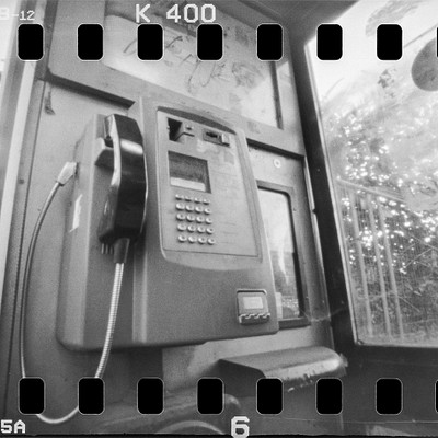 The image features an old-fashioned payphone with a black and white color scheme. It is placed in front of a window, possibly at a bus stop or a public location. The phone has a dial pad on the side for making calls, and it appears to be outdated compared to modern smartphones.