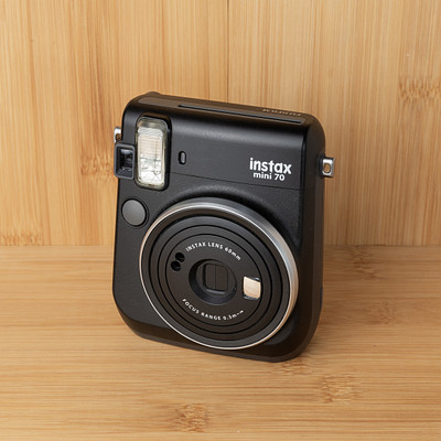 The image features a black Instax camera sitting on top of a wooden table. The camera is placed in the center of the scene, drawing attention to its unique design and function. The wooden surface provides an appealing backdrop for this modern photography device.