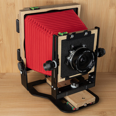 The image features a wooden box with an old-fashioned camera inside. The camera is red and black, giving it a vintage appearance. It appears to be a large format camera, possibly used for photography or filming purposes.
