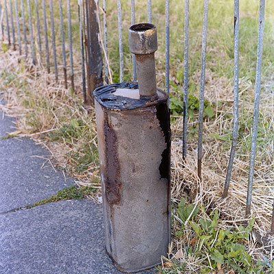 The image features a metal pipe or barrel sitting on the sidewalk next to a chain-link fence. It appears to be an old, rusted container that has been placed there for some time. The fence is located behind the object and extends across the scene.