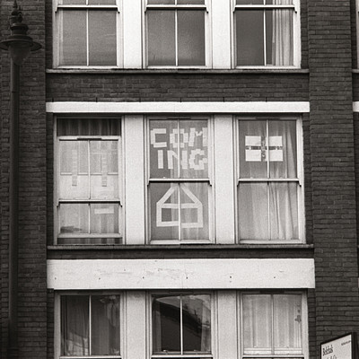 The image is a black and white photo of an old building with many windows. There are several signs on the windows, including one that says "coming." The building has a brick facade, giving it an aged appearance. The windows have curtains covering them, adding to the vintage feel of the scene.