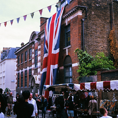 The image depicts a busy street scene with people walking around and engaging in various activities. There are several individuals scattered throughout the scene, some closer to the foreground while others are further away. A large British flag is hanging above the street, adding a sense of national pride to the atmosphere.