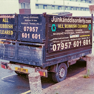 The image is a black and white photo of an old truck parked on the side of a street. The truck has a sign on its back, advertising "Junkand Disorderly" or "All Rubbish Clear." There are several cars in the background, including one behind the truck and others further down the road. A stop sign can be seen near the top right corner of the image.