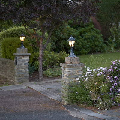 The image features a beautifully landscaped driveway with a cobblestone path leading to a house. Along the path, there are two street lamps positioned at different points, providing ample lighting for the area. In addition to the lamps, there is also a bench situated near the entrance of the property.