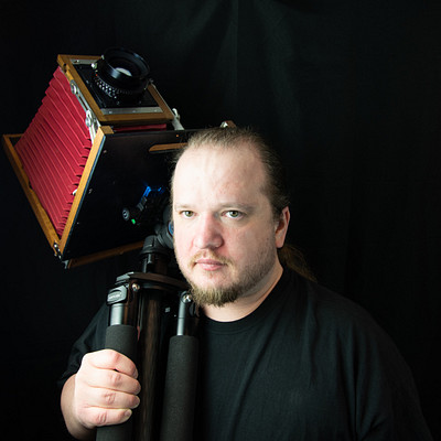 The image features a man wearing a black shirt and holding a camera in his hands. He is standing in front of a dark background, possibly for a photo shoot or filming session. The man appears to be focused on the task at hand, as he holds the camera with both hands.