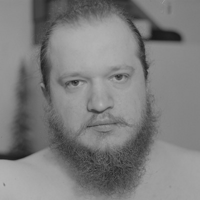 The image is a black and white photo of a man with a beard. He has long hair, which appears to be brown in color. The man's facial features are prominent, including his eyes, nose, mouth, and mustache.