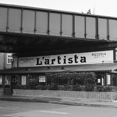 The image is a black and white photo of an outdoor pizza restaurant called La Artista. The restaurant has a brick facade, and there are several potted plants placed around the area. A large sign hangs above the entrance to the restaurant, indicating its name.