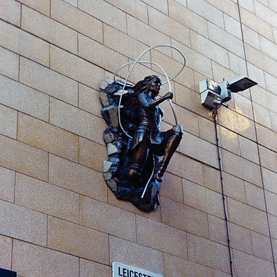 The image features a statue of a man holding a guitar, mounted on the side of a building. The statue is positioned above a street sign that reads "Leicester Court WC2." In addition to the statue and sign, there are two traffic lights visible in the scene, one located near the top right corner and another further down towards the bottom left.