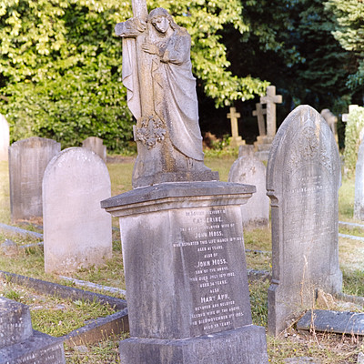 The image features a cemetery with several headstones and monuments. A prominent statue of a person, possibly a saint or religious figure, is standing on top of one of the graves. This statue appears to be made of stone and has an intricate design.