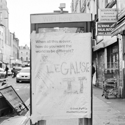 The image is a black and white photo of a city street with a telephone booth in the foreground. A sign on the phone booth reads "Legalise It." There are several people walking around, some closer to the camera while others are further away. In addition to pedestrians, there are multiple cars parked or driving along the street.