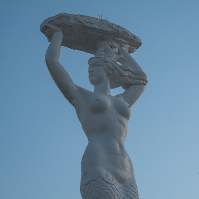 The image features a statue of a woman holding a large tray or basket on her head. She appears to be a mermaid, as she is depicted with a fish-like tail. The statue is positioned outdoors in front of a blue sky, giving it an artistic and serene atmosphere.