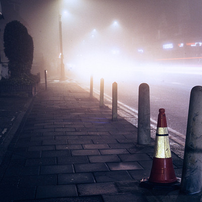 The image depicts a foggy city street at night, with a yellow traffic cone sitting on the sidewalk next to a pole. There are several cars parked or driving along the road, and some of them appear blurry due to the foggy conditions. A few people can be seen walking around in the area, adding to the overall atmosphere of the scene.
