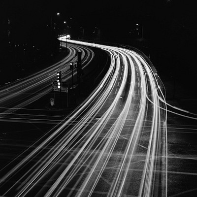 The image is a black and white photo of a highway at night, with traffic moving in both directions. There are multiple cars visible on the road, some closer to the foreground while others are further away. A few traffic lights can be seen along the highway, helping to control the flow of vehicles.