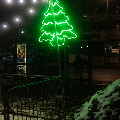 The image features a green neon Christmas tree with lights on it, standing in the snow. It is located near a fence and appears to be lit up at night. There are also two people visible in the scene, one closer to the left side of the frame and another further back towards the right.