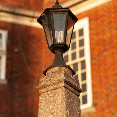 The image features a lamp post with a light on it, situated in front of a red brick building. The lamp post is made of stone and has a black top. The building appears to be an old-fashioned structure with a window on the side.