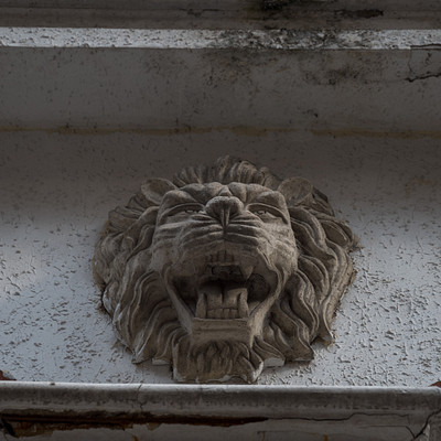 The image features a white wall with an ornate lion head sculpture. The lion's mouth is open, giving the impression that it is roaring or making a sound. The sculpture appears to be made of stone and has intricate details, adding character to the wall.