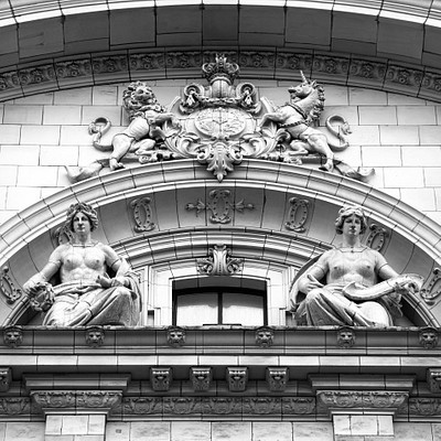 The image is a black and white photo of an ornate building with statues on the front. There are two prominent statues, one on each side of the building, depicting women sitting in chairs. These statues appear to be part of a larger decorative design or architectural feature.