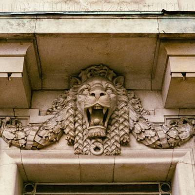The image features a building with an ornate design on its facade. A prominent lion statue is the centerpiece of this decoration, with its mouth wide open and teeth bared in a fierce expression. The lion's head is positioned above a doorway, adding to the grandeur of the architectural details.