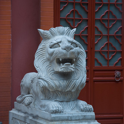 The image features a large stone lion statue, which is situated on top of a pedestal. The lion appears to be roaring and has its mouth open wide, giving the impression that it's yawning or making a funny face. The statue is positioned in front of a red building with an ornate design, adding a striking contrast between the stone lion and the colorful background.