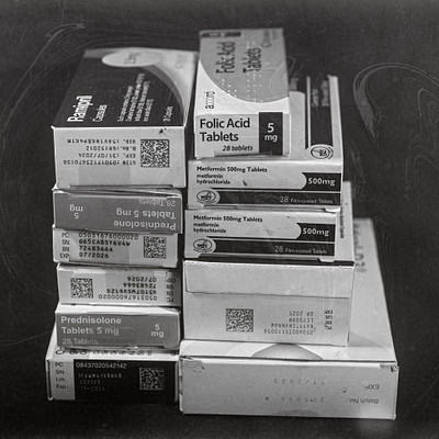 The image is a black and white photo of several boxes stacked on top of each other. These boxes are filled with medications, including pills and tablets. There are at least 13 different types of medication visible in the image, arranged in various positions within the stacks.